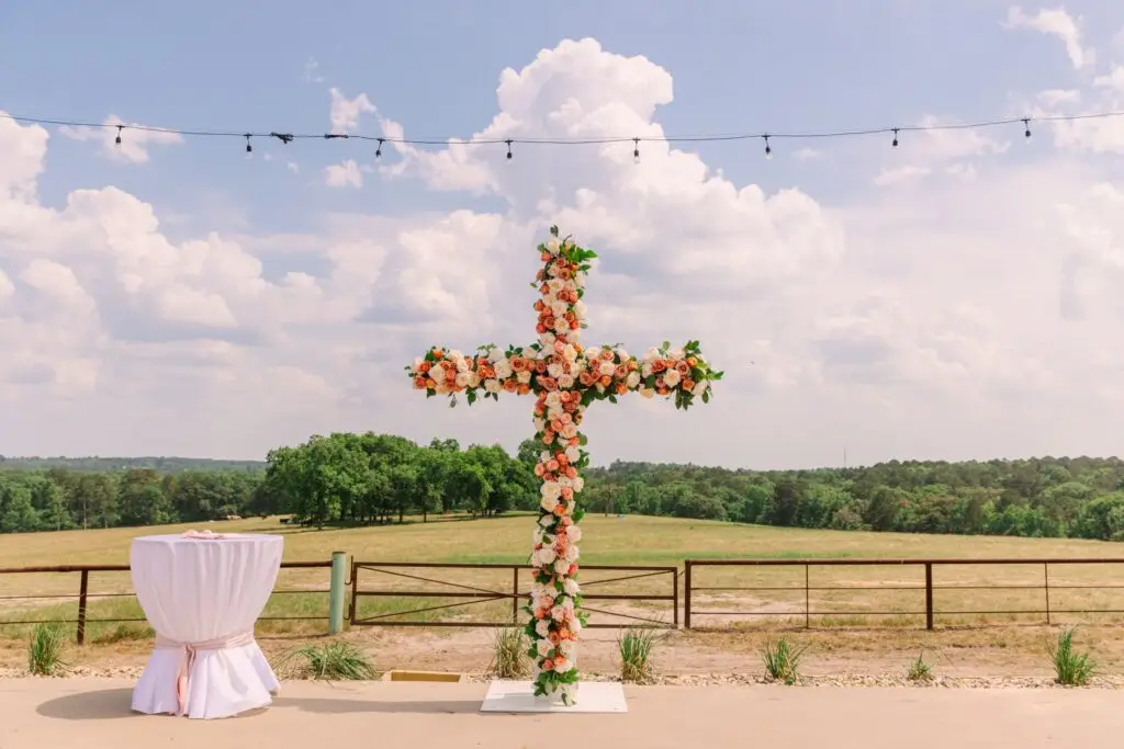 An outdoor wedding setup at El Cerrito with a floral cross, string lights, and a white-clothed table beneath the sunny sky.