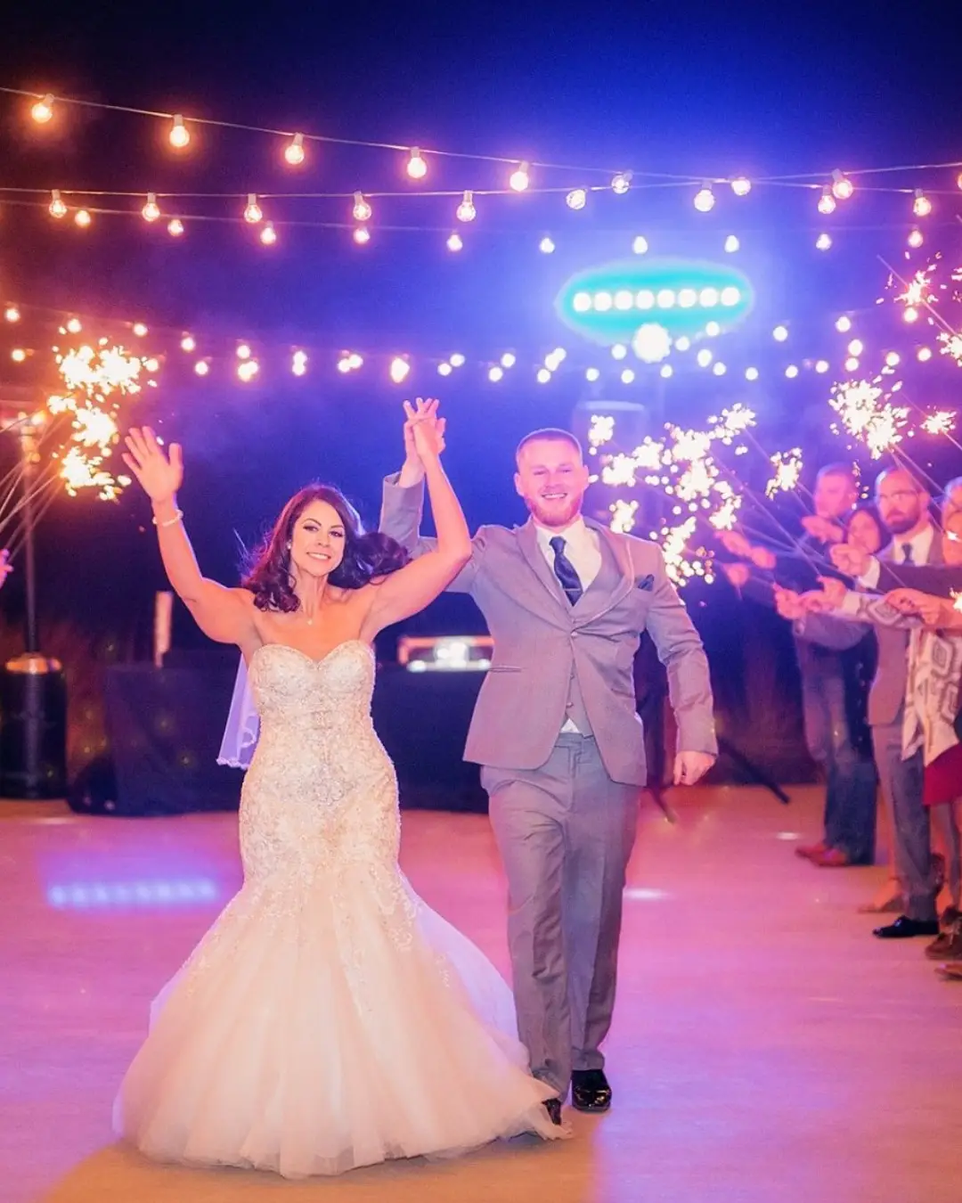 At El Cerrito, a joyful bride and groom walk hand in hand beneath glowing string lights as guests celebrate with sparklers at night.