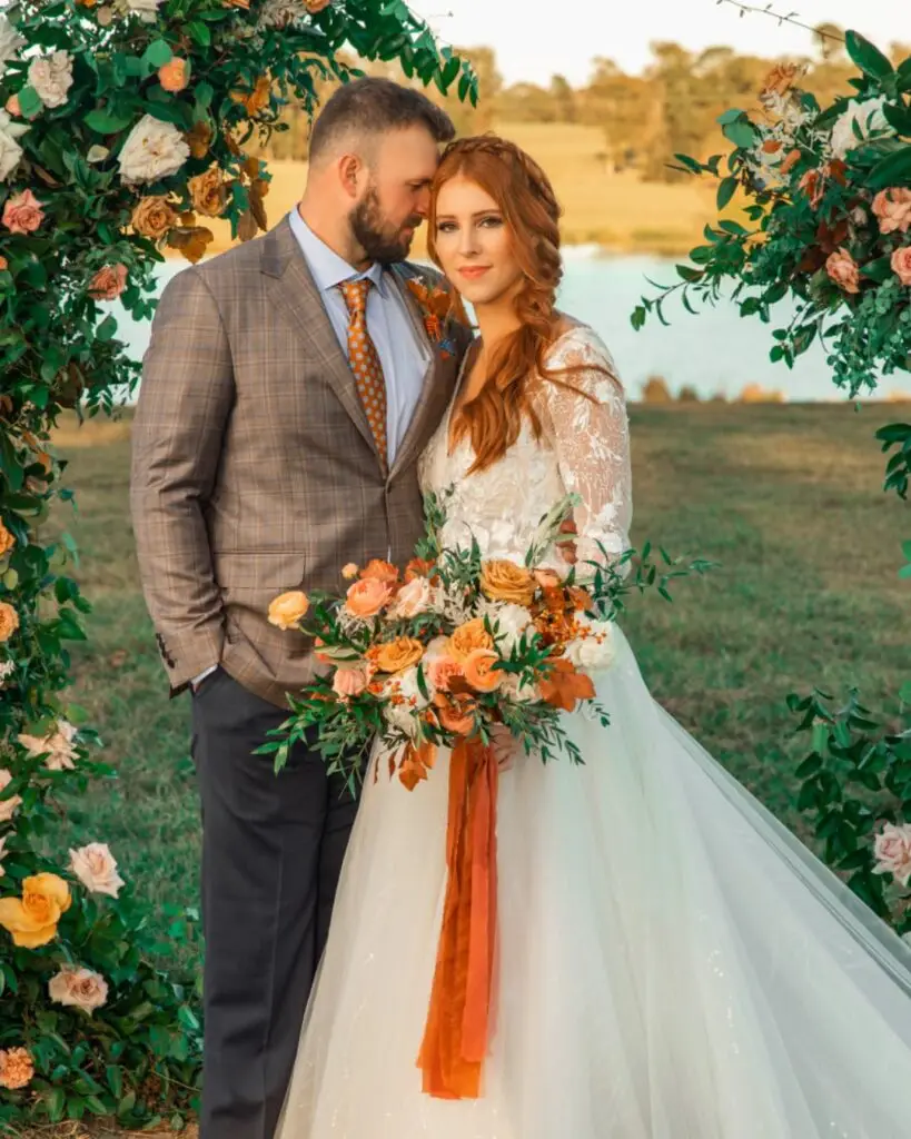 An elegant bride and groom stand beneath a floral arch by the lake at El Cerrito, creating a romantic outdoor wedding scene.